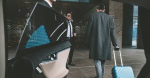 Someone holds a door open to a vehicle. A man carrying a blue suitcase walks toward another man in business attire.