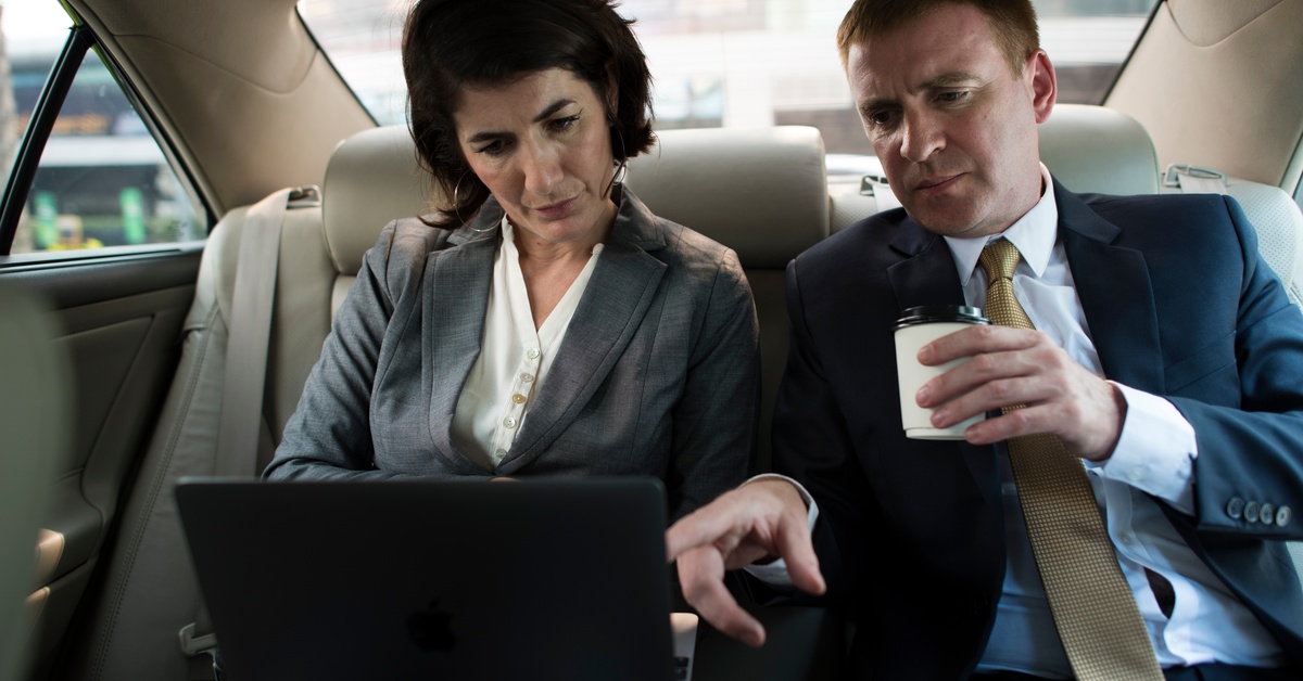 A man and woman in business professional clothing sit in the back seat of a car. They both look at a laptop open on the woman's legs.