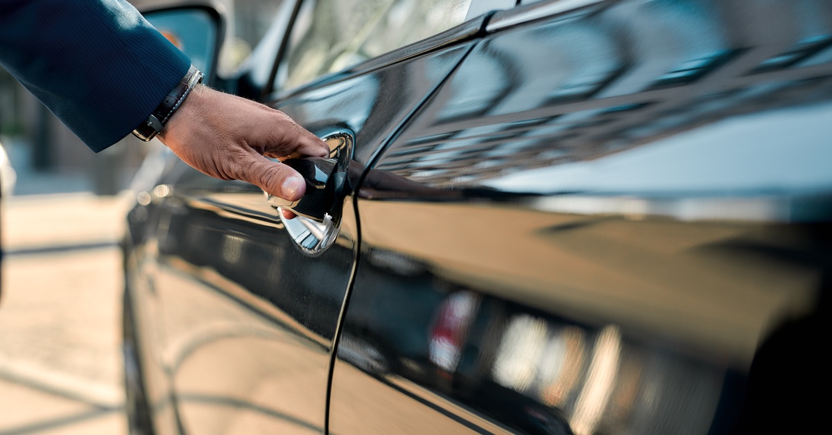A close-up of someone's hand on the handle of a black car door. The car is outdoors with the sun shining.