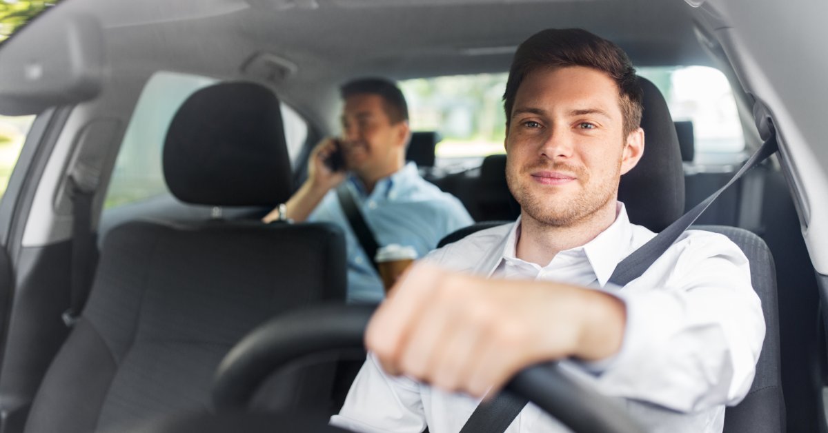 A man wearing a white shirt sits behind the wheel of a car. Another man with his phone against his ear sits in the backseat.