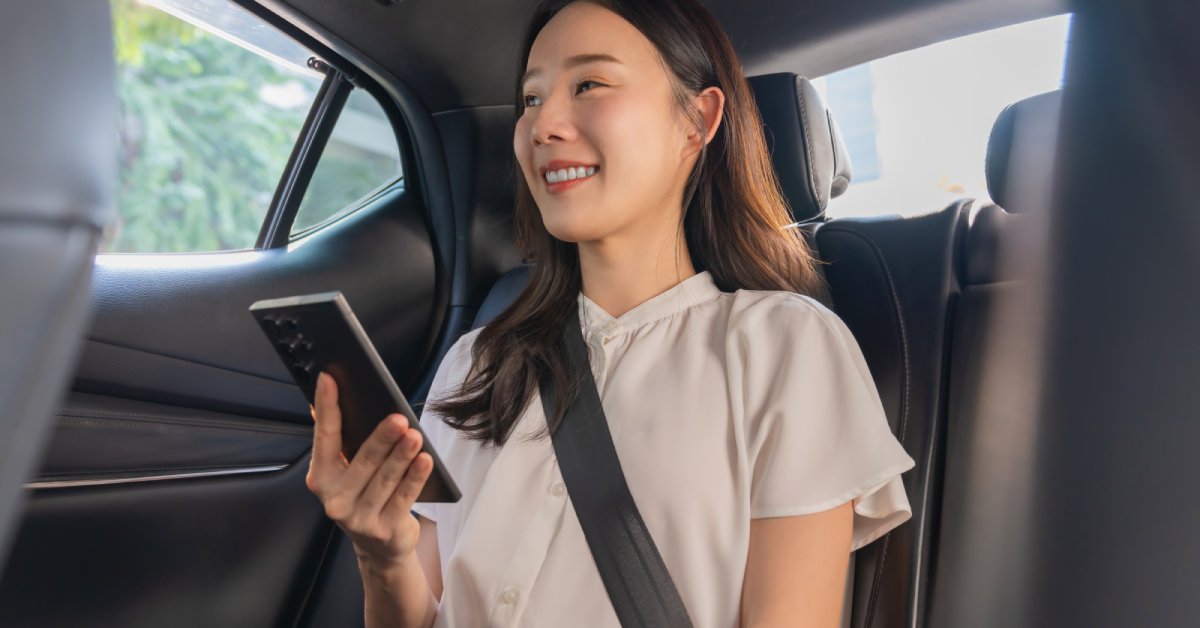 A woman sits in the back seat of a car and holds her phone up in front of her. The seats are black leather.