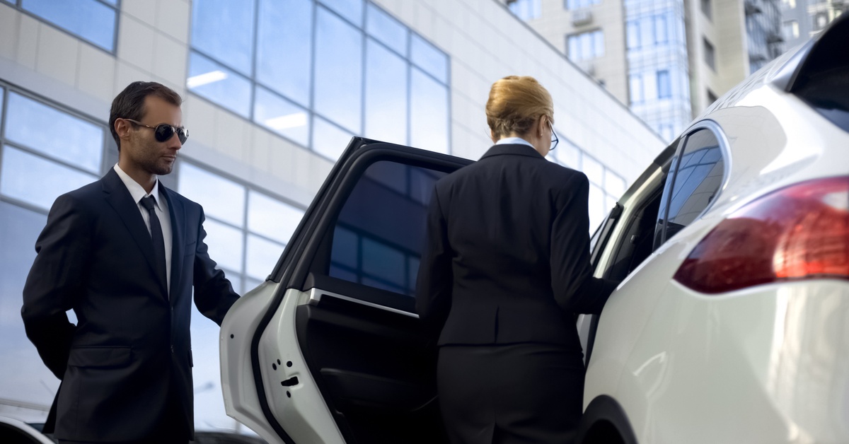 A man wearing a suit and sunglasses holds open the back door of an SUV for a woman in business clothing.