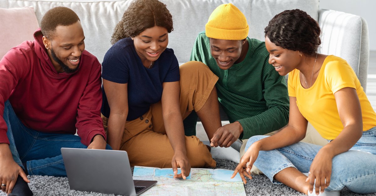 A group of African American friends sits on a carpeted floor. They are looking at a map and a laptop together.