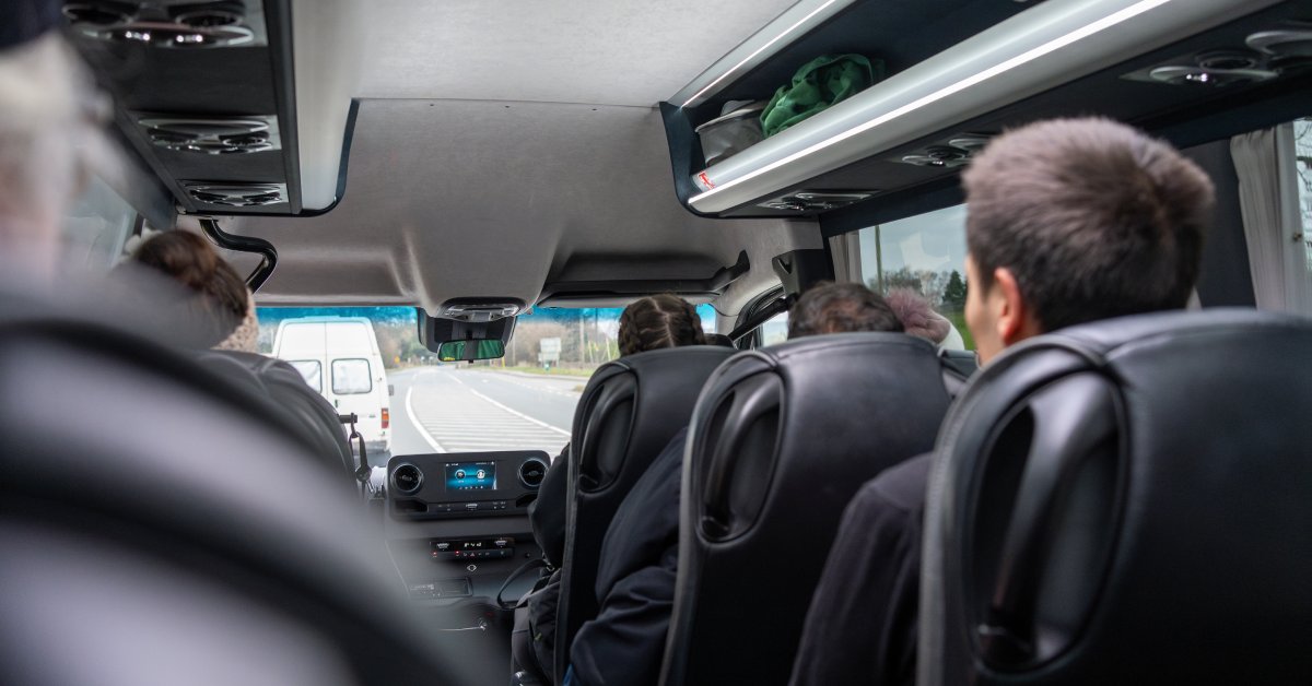 The inside of a mini shuttle bus with black leather interior and overhead storage. The backs of people's heads are visible.
