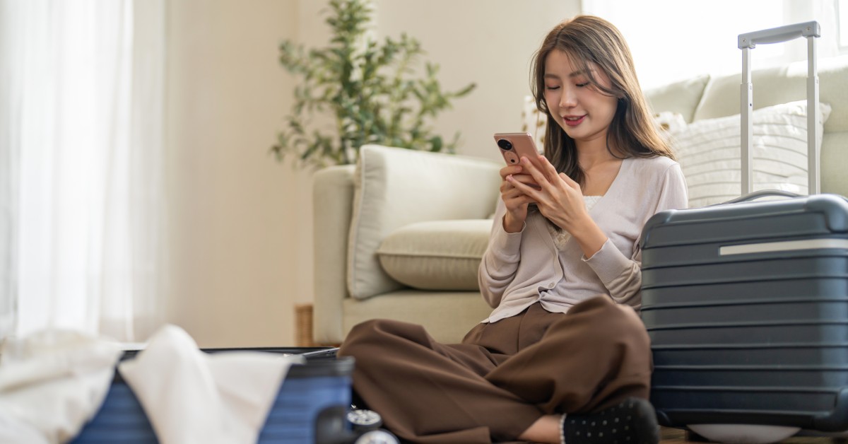 A woman sits on the floor with her back against a couch. She looks at her phone with her luggage next to her.