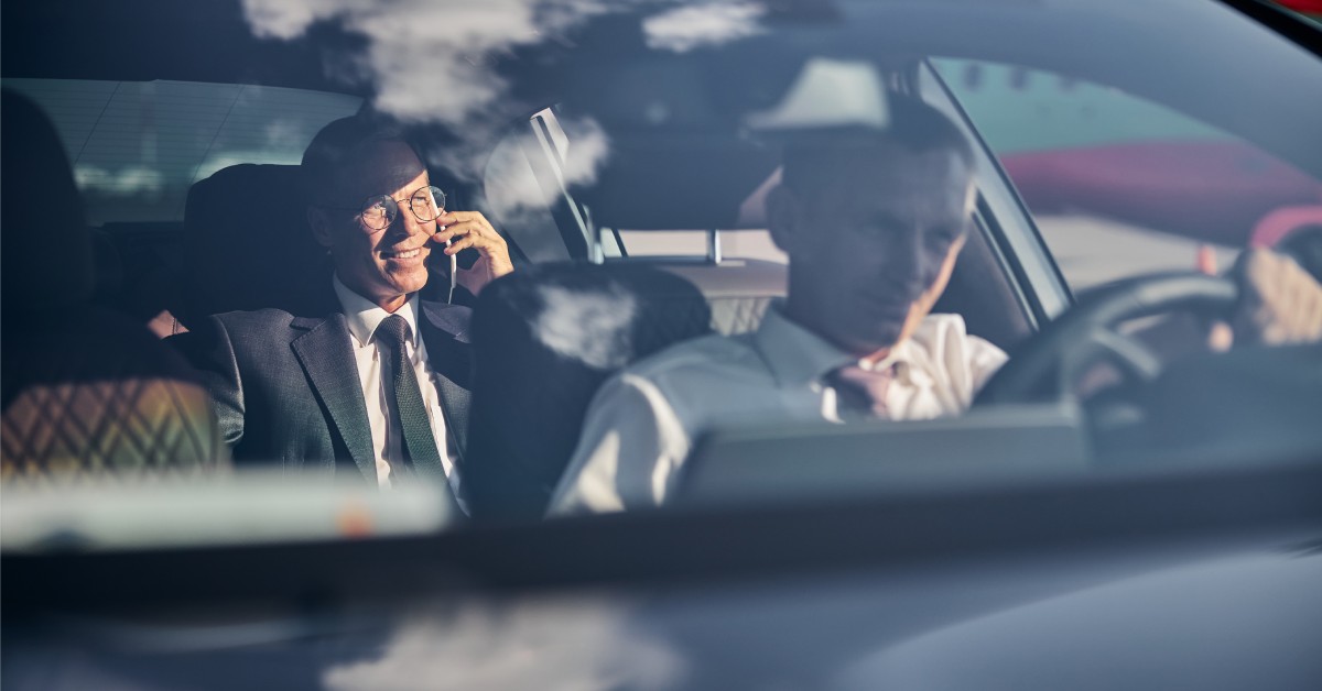 A man in a suit talks on the phone in the back seat of a car. A driver is focused ahead with clouds reflected on the windshield.